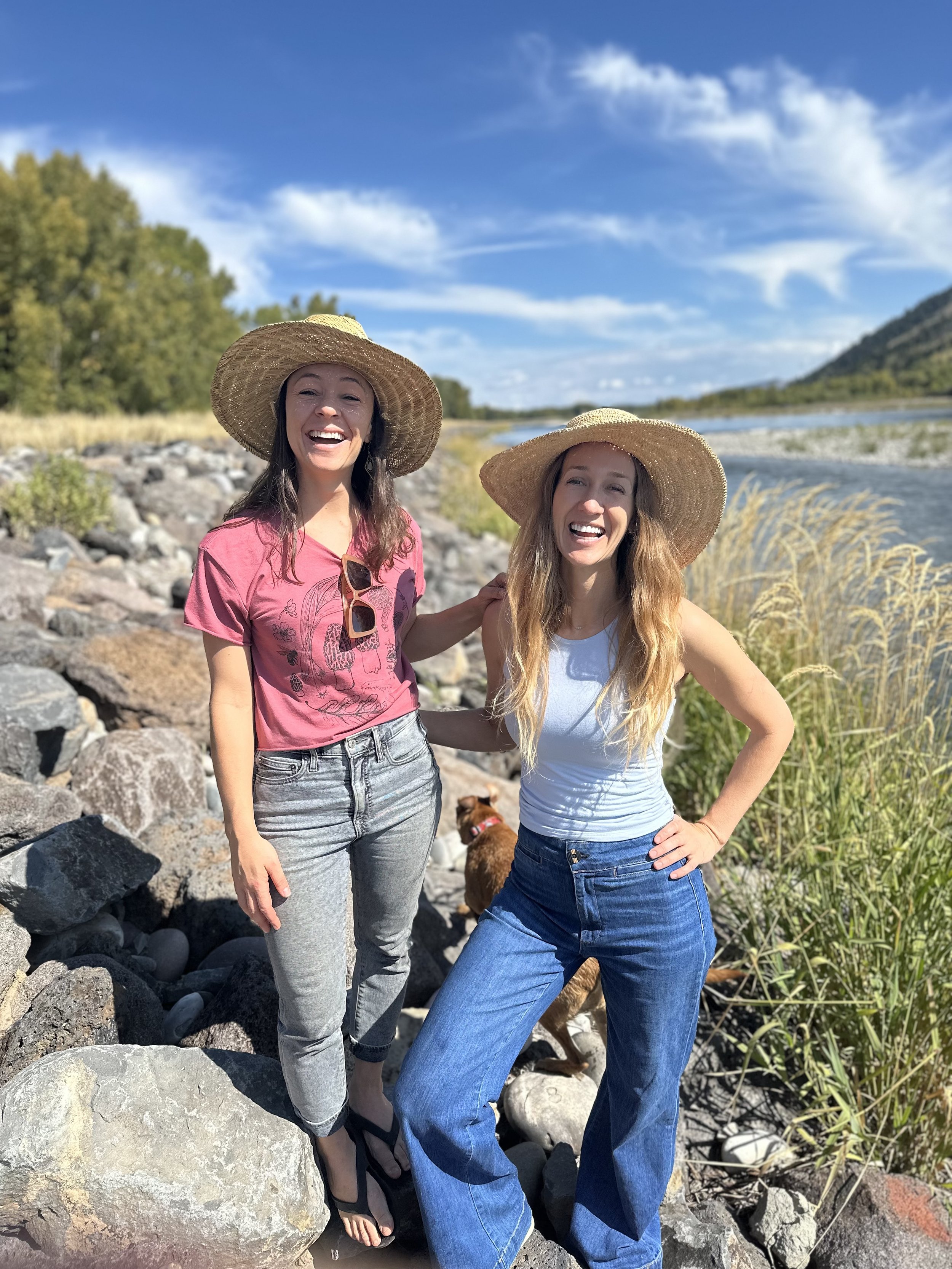 Dr. Kelly Baxter and Dr. Kelsey Sherman by a river in straw hats, laughing in golden afternoon light